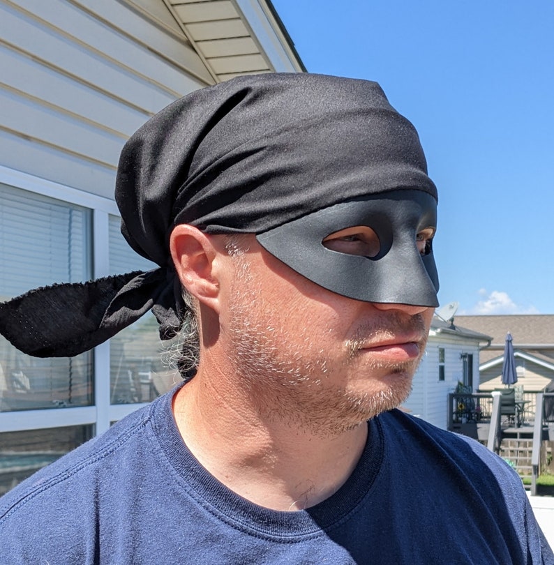 May include: A person wearing a black bandana and a black eye mask. The bandana is tied around the person's head and the eye mask covers the person's eyes.