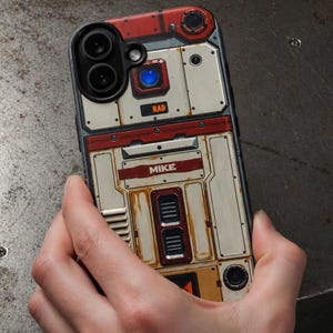 May include: A phone case featuring a weathered, industrial-style design with off-white and red panels, metallic accents, and a blue circular light. The case displays the text "RAD" and "MIKE" in bold lettering, mimicking a mechanical, sci-fi aesthetic.