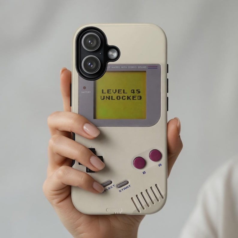 May include: A beige smartphone case designed to resemble a vintage handheld gaming console. The screen area displays the text LEVEL 45 UNLOCKED in a pixelated font. It features two purple circular buttons, two grey oval buttons, and a speaker grill.