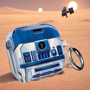 May include: A blue and white protective case for wireless earbuds, designed to resemble a robot. The case has a silver carabiner clip attached. The design includes rectangular and circular details, with a small orange accent.