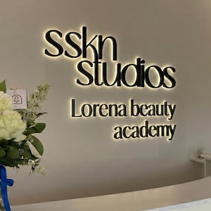 以下が含まれることがあります： 壁に取り付けられた看板には、黒い文字でsskn studiosとLorena beauty academyと記されており、背後から光を放つバックライト効果があります。文字は明るい色の壁に設置され、各文字の周囲に柔らかな光の輪を作り出しています。