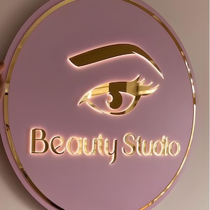 Puede incluir: Un letrero redondo rosa de Beauty Studio con borde y letras doradas. El letrero presenta un diseño de ojo dorado y las palabras "Beauty Studio" en una fuente dorada brillante. El letrero está sostenido por una persona.