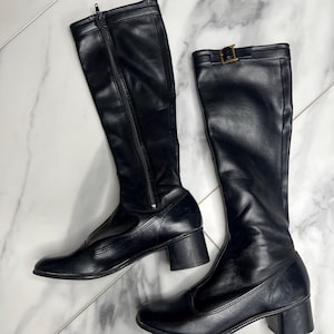 Puede incluir: Un par de botas negras hasta la rodilla con acabado liso, tacón de bloque, punta redondeada y cierre de cremallera lateral. Una de las botas incluye una tira decorativa cerca de la parte superior con una hebilla de metal en tono dorado.