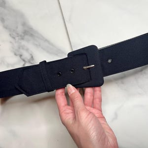 May include: A wide, black belt with a large square buckle and a silver-colored pin. The belt is held by a hand, showcasing its texture and design. The belt has two eyelets for adjustment.