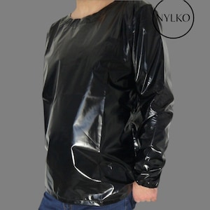 Può includere: Camicia nera a maniche lunghe con una superficie lucida e riflettente. La camicia ha un girocollo e maniche lunghe. Il marchio "NYLKO" è in un cerchio in alto a destra. La camicia è indossata con jeans blu.