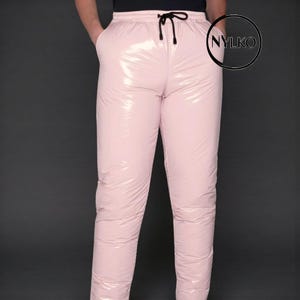Puede incluir: Pantalones acolchados rosa claro brillante con cintura con cordón y bolsillos. Los pantalones tienen un acabado brillante y reflectante y un ajuste relajado. Los pantalones se muestran sobre un fondo gris oscuro. La marca "NYLKO" es visible en un círculo en el lado derecho.