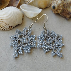 May include: A pair of pale blue snowflake earrings with silver hooks. The earrings are crafted from delicate lace and tiny beads, featuring an intricate, openwork design. The background includes seashells.