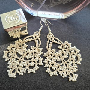 May include: A pair of cream-colored, handmade lace earrings with small white beads. The earrings are displayed on a dark surface, with a perfume bottle and a silver compact in the background.