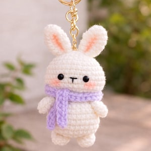 May include: A handmade white crochet bunny keychain with a gold-colored ring. The bunny has black eyes, pink cheeks, and a purple scarf. The bunny's ears have pink inner details.