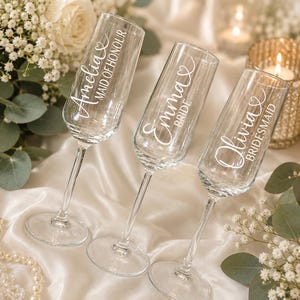 May include: Three clear glass champagne flutes with white lettering. The first glass reads Amelia Maid of Honour, the second reads Emma Bride, and the third reads Olivia Bridesmaid. Each glass features a small heart icon above the name.