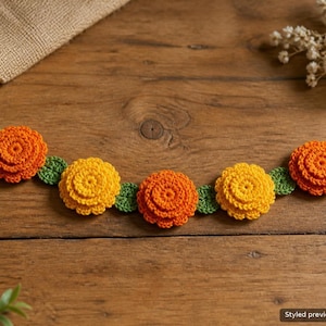 May include: A crochet garland featuring five layered flowers in alternating shades of orange and yellow, each accented with small green leaves. The flowers are connected by a thin green crocheted cord, creating a continuous floral strand.