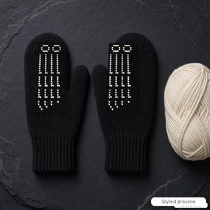 Può includere: Un paio di muffole in maglia nera con un motivo grafico bianco di linee verticali e piccole forme ovali sul dorso della mano, accompagnate da un gomitolo di lana color crema accanto alle muffole su una superficie scura e strutturata.