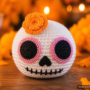 Könnte beinhalten: Ein runder, weiß gehäkelter Totenkopf mit strukturierter Oberfläche. Er hat große, kreisförmige schwarze Augenhöhlen, die mit rosa Garn umrandet sind, eine kleine schwarze dreieckige Nase und einen aufgestickten schwarzen Mund.