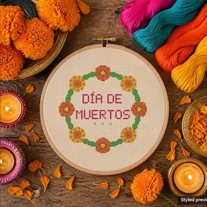 May include: A wooden embroidery hoop containing a cross-stitch pattern with the text DÍA DE MUERTOS in pink, surrounded by a circular wreath of orange and yellow flowers, accompanied by loose marigold flowers and colorful yarn.