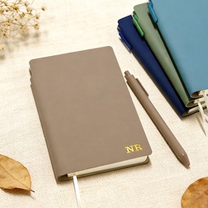 May include: A collection of notebooks in various colors, including taupe, blue, and green, with matching pens. The taupe notebook has the gold letters "NR" on the cover. Each notebook has a ribbon bookmark.