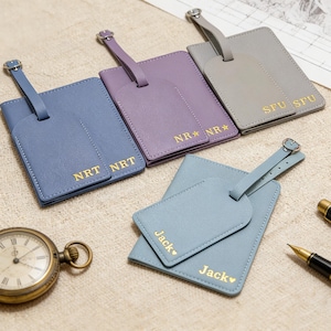 Puede incluir: Una colección de etiquetas de equipaje personalizadas en varios colores: azul, morado y gris. Cada etiqueta presenta un diseño rectangular con una etiqueta más pequeña adjunta y letras doradas con nombres e iniciales. También se ve un reloj de bolsillo y un bolígrafo vintage.