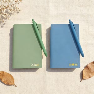 Może przedstawiać: Dwa notatniki, jeden w kolorze szałwii, drugi w kolorze niebieskim, każdy z pasującym długopisem. Zielony notatnik ma imię "Alice" złotymi literami, a niebieski notatnik ma "B ♡ M". Długopisy spoczywają na notatnikach.