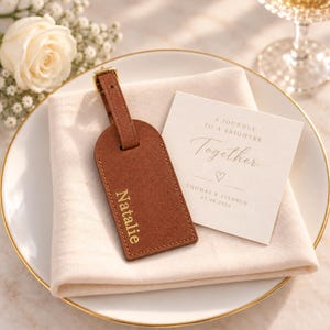 May include: A brown luggage tag with the name Natalie embossed in gold, featuring a matching strap and buckle. Beside it lies a square white card with the text A JOURNEY TO A BRIGHTER Together, a heart icon, and the names THOMAS & CULHWCH 07.08.2026.