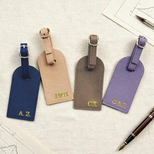 May include: Four personalized luggage tags in navy blue, tan, brown, and lavender. Each tag has a gold buckle and strap, with gold initials. The tags are arranged on a textured white surface, with a pen and map in the background.