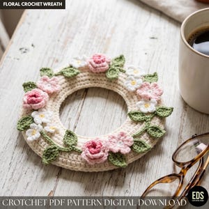 May include: A cream-colored, crocheted floral wreath adorned with pink and white flowers and green leaves. The wreath is circular and rests on a light-colored wooden surface. Text at the top reads "FLORAL CROCHET WREATH". The bottom text reads "CROCHET PDF PATTERN DIGITAL DOWNLOAD".