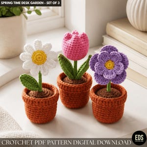 Può includere: Tre fiori all'uncinetto in vasi di terracotta. I fiori includono una margherita bianca con centro giallo, un tulipano rosa e un fiore viola con centro giallo. Il testo "SPRING TIME DESK GARDEN - SET OF 3" è in alto.