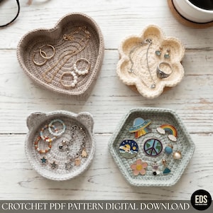 May include: Four crocheted trinket trays in heart, flower, cat-ear, and hexagonal shapes. The trays hold jewelry, including gold hoops, pearl rings, beaded bracelets, and colorful enamel pins featuring a UFO, rainbow, and peace sign.