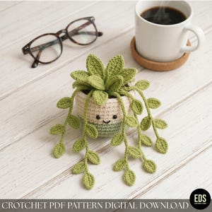 Puede incluir: Una planta de ganchillo en una maceta crema y verde con una cara sonriente. La planta tiene hojas y enredaderas verdes. La imagen también incluye un par de gafas y una taza de café.