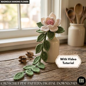 May include: A crocheted magnolia flower with a pink center and off-white petals sits in a small white vase. Green crocheted leaves and a stem extend from the flower. The text "Magnolia Hanging Flower" and "With Video Tutorial" are visible.