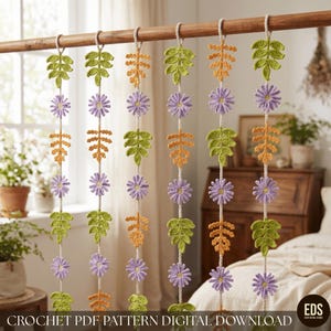 May include: Six vertical hanging garlands made of crocheted elements suspended from a wooden rod. Each garland features alternating light purple daisy-like flowers, bright green fern-like leaves, and orange fern-like leaves on a white cord.