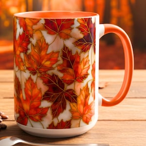 May include: A ceramic mug with an orange handle and interior, decorated with a pattern of autumn leaves in shades of red, orange, and brown. The mug sits on a wooden surface with a macaron, coffee beans, and a spoon.