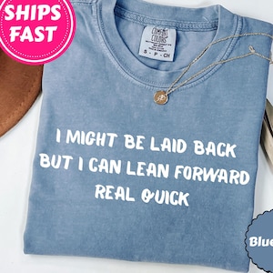 以下が含まれることがあります： 「I MIGHT BE LAID BACK BUT I CAN LEAN FORWARD REAL QUICK」の白文字がプリントされた、ブルーデニムカラーのTシャツ。ゴールドのネックレスと茶色の帽子も写っています。
