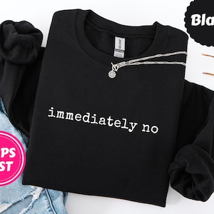 以下が含まれることがあります： 「immediately no」の白文字が入った黒のクルーネックトレーナー。円形のペンダントが付いたシルバーのネックレスが前面にかけられています。トレーナーは袖を内側に折りたたんであります。