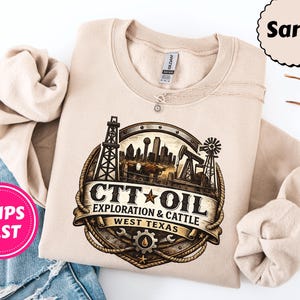 Może przedstawiać: Beżowa bluza z obszerną grafiką. Projekt przedstawia wieże wiertnicze, krajobraz miejski i wiatrak, z napisem "CTT OIL EXPLORATION & CATTLE WEST TEXAS" w okrągłym emblemacie.