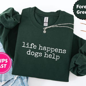 May include: Forest green sweatshirt with the text "life happens dogs help" in white. A silver necklace with a pendant is draped across the front. The sweatshirt is folded on top of a pair of blue jeans.