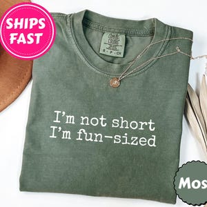 May include: A moss green t-shirt with the text "I'm not short I'm fun-sized" in white. A gold necklace with a pendant rests on the shirt. The shirt is folded, with a brown hat and dried leaves in the background.