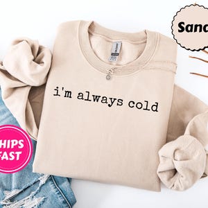 Peut inclure: Un sweat-shirt beige avec l'inscription "i'm always cold" imprim&eacute;e en noir. Le sweat-shirt est pli&eacute;, avec un collier argent&eacute; visible. Un autocollant rose avec "SHIPS FAST" est dans le coin inf&eacute;rieur gauche. Le mot "Sand" est dans un cercle.