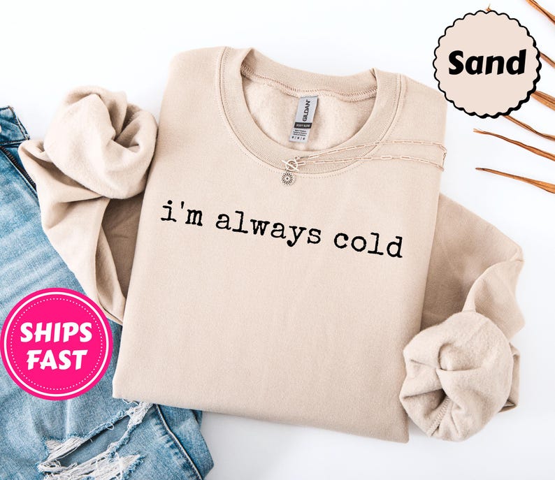 Peut inclure: Un sweat-shirt beige avec l'inscription "i'm always cold" imprim&eacute;e en noir. Le sweat-shirt est pli&eacute;, avec un collier argent&eacute; visible. Un autocollant rose avec "SHIPS FAST" est dans le coin inf&eacute;rieur gauche. Le mot "Sand" est dans un cercle.