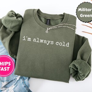 Peut inclure: Sweat-shirt vert militaire avec l'inscription blanche "i'm always cold". Le sweat-shirt &agrave; col rond et &agrave; manches longues est orn&eacute; d'un collier argent&eacute; et pli&eacute; sur un jean bleu.