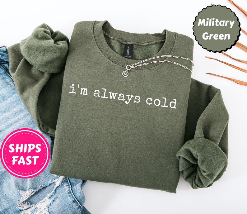 Peut inclure: Sweat-shirt vert militaire avec l'inscription blanche "i'm always cold". Le sweat-shirt &agrave; col rond et &agrave; manches longues est orn&eacute; d'un collier argent&eacute; et pli&eacute; sur un jean bleu.