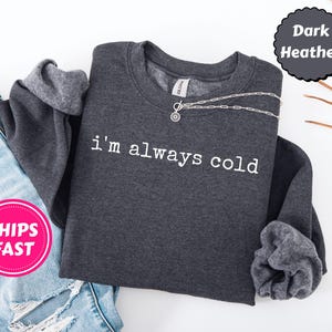 Peut inclure: Sweat-shirt gris fonc&eacute; avec l'inscription blanche &laquo;&nbsp;i'm always cold&nbsp;&raquo;. Un collier argent&eacute; avec un pendentif circulaire est pos&eacute; sur le sweat-shirt. Le sweat-shirt est &agrave; c&ocirc;t&eacute; d'un jean bleu.