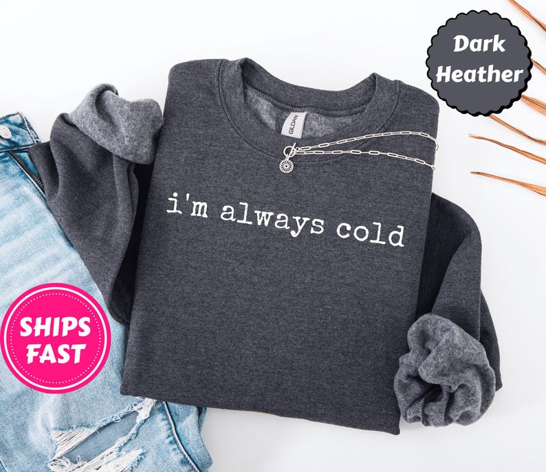 Peut inclure: Sweat-shirt gris fonc&eacute; avec l'inscription blanche &laquo;&nbsp;i'm always cold&nbsp;&raquo;. Un collier argent&eacute; avec un pendentif circulaire est pos&eacute; sur le sweat-shirt. Le sweat-shirt est &agrave; c&ocirc;t&eacute; d'un jean bleu.