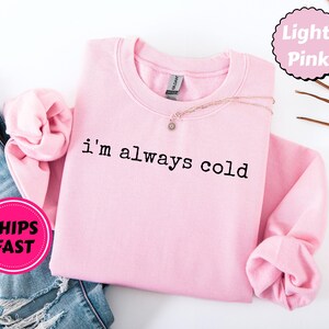 Peut inclure: Sweat-shirt rose clair avec l'inscription noire "I'm always cold". Les manches sont repli&eacute;es. Un collier avec un petit pendentif est pos&eacute; sur le v&ecirc;tement. Les mots "Light Pink" sont dans un cercle rose.