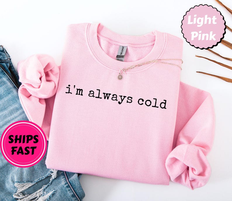 Peut inclure: Sweat-shirt rose clair avec l'inscription noire "I'm always cold". Les manches sont repli&eacute;es. Un collier avec un petit pendentif est pos&eacute; sur le v&ecirc;tement. Les mots "Light Pink" sont dans un cercle rose.
