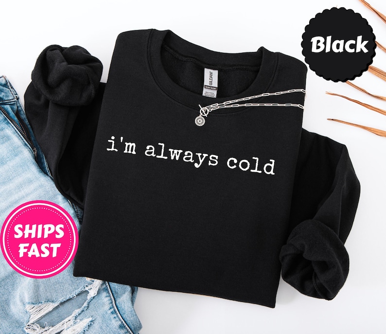 Peut inclure: Sweat-shirt noir &agrave; col rond avec le texte "i'm always cold" en blanc. Un collier argent&eacute; avec un pendentif circulaire repose sur le sweat-shirt. Le sweat-shirt est pli&eacute;, avec les manches retrouss&eacute;es. Le mot "Black" est dans un cercle noir.
