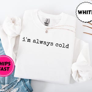 Peut inclure: Sweat-shirt blanc &agrave; col rond avec le texte "i'm always cold" en noir. Un collier argent&eacute; avec un petit pendentif repose sur le sweat-shirt. Un cercle rose avec les mots "SHIPS FAST" est visible.