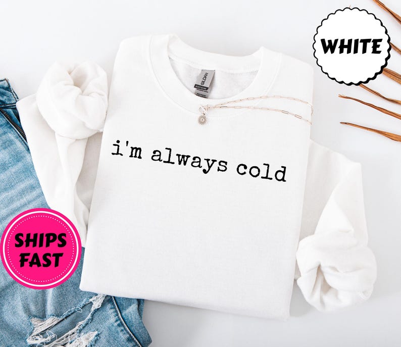 Peut inclure: Sweat-shirt blanc &agrave; col rond avec le texte "i'm always cold" en noir. Un collier argent&eacute; avec un petit pendentif repose sur le sweat-shirt. Un cercle rose avec les mots "SHIPS FAST" est visible.
