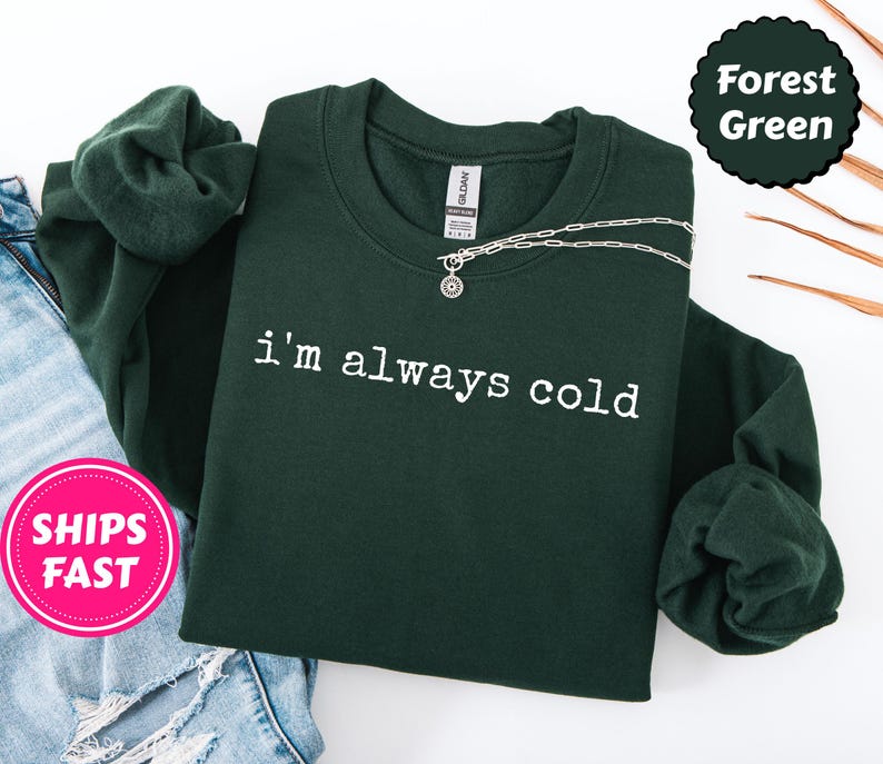 Peut inclure: Sweat-shirt vert for&ecirc;t avec l'inscription blanche "I'm always cold". Un collier argent&eacute; avec un pendentif est pos&eacute; sur le sweat-shirt. Le sweat-shirt est pli&eacute; &agrave; c&ocirc;t&eacute; d'un jean bleu.