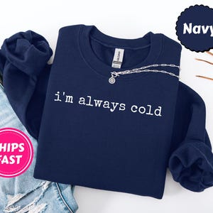 Peut inclure: Un sweat-shirt bleu marine avec le texte blanc "I'm always cold". Un collier argent&eacute; avec un pendentif est drap&eacute; sur le devant. Le sweat-shirt est pli&eacute; sur un jean bleu.