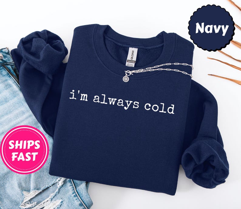 Peut inclure: Un sweat-shirt bleu marine avec le texte blanc "I'm always cold". Un collier argent&eacute; avec un pendentif est drap&eacute; sur le devant. Le sweat-shirt est pli&eacute; sur un jean bleu.