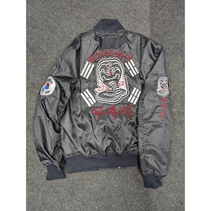 Puede incluir: Chaqueta bomber de satén negro con el logo de Cobra Kai bordado en la espalda, con una cobra y texto en blanco y rojo. Incluye parches con la bandera coreana y el emblema de Cobra Kai. Puños y cuello de canalé.
